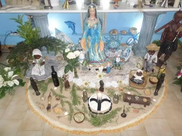 Altar de Iemanjá com a imagem da orixá ao centro, conchas, ervas, flores brancas e velas, rodeado pelas imagens dos pretos velhos