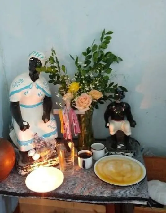 Firmeza para os pretos velhos com as imagens de uma preta velha e de um preto velho, café, rosas, vela acesa e um prato de oferenda