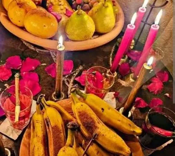 Oferenda com bananas, peras, laranjas, pétalas de rosa, taças e velas rosa e douradas acesas