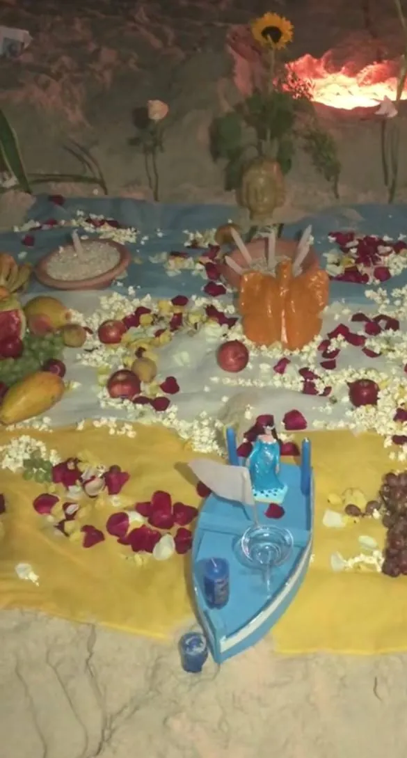 Oferenda para Iemanjá na areia da praia, com barco azul, imagem da orixá, frutas, pipoca, pétalas de rosa e velas