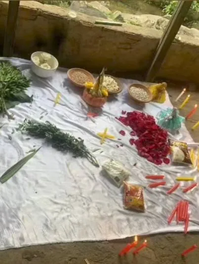 Oferenda sobre pano branco com ervas, pétalas de rosa, tigelas de grãos, flores amarelas e velas coloridas acesas